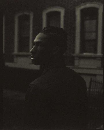 Monochrome portrait of a stylish adult male with locs, gazing thoughtfully with an architectural backdrop featuring framed windows.