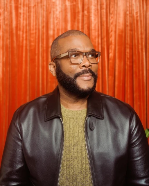 Portrait of African-American man, Tyler Perry, in leather jacket and glasses, looking off-camera, against an orange curtain backdrop.