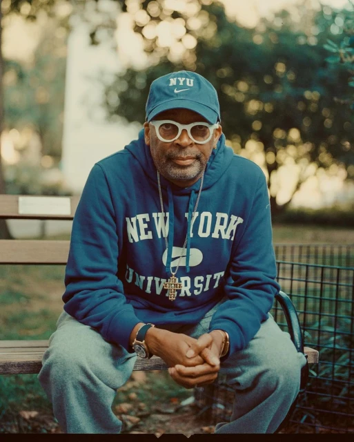 Portrait of Spike Lee sitting on a park bench wearing a blue NYU hoodie, hat, and white glasses in an outdoor setting.