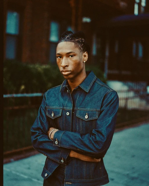 Young adult in a denim jacket with arms crossed stands on a city sidewalk near a brick building. Medium shot, eye-level perspective.