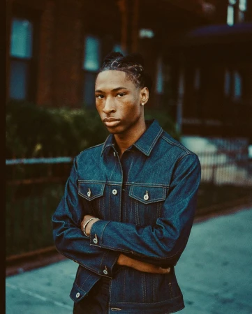 Young adult in a denim jacket with arms crossed stands on a city sidewalk near a brick building. Medium shot, eye-level perspective.