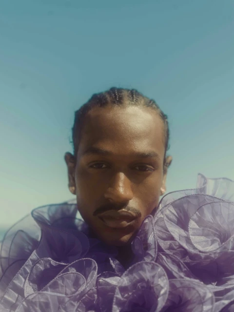 Close-up portrait of a Black male model with cornrow braids and a mustache, wearing a purple ruffled top, against a light blue sky.