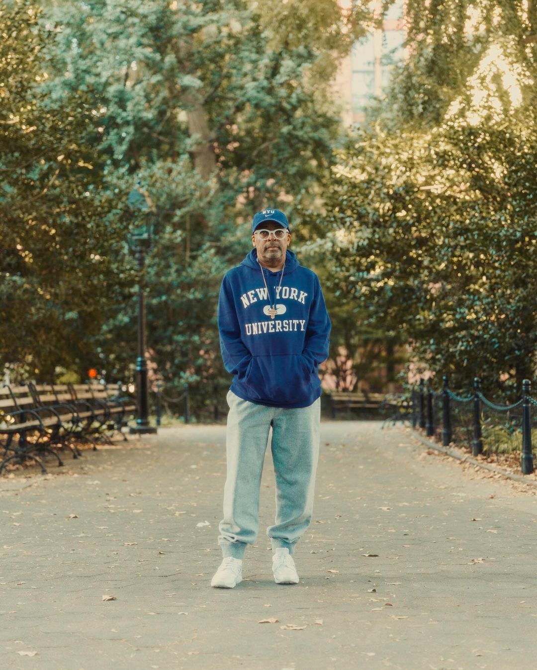 Adult male stands on a park path wearing a blue New York University hoodie, grey sweatpants, and white sneakers surrounded by trees.