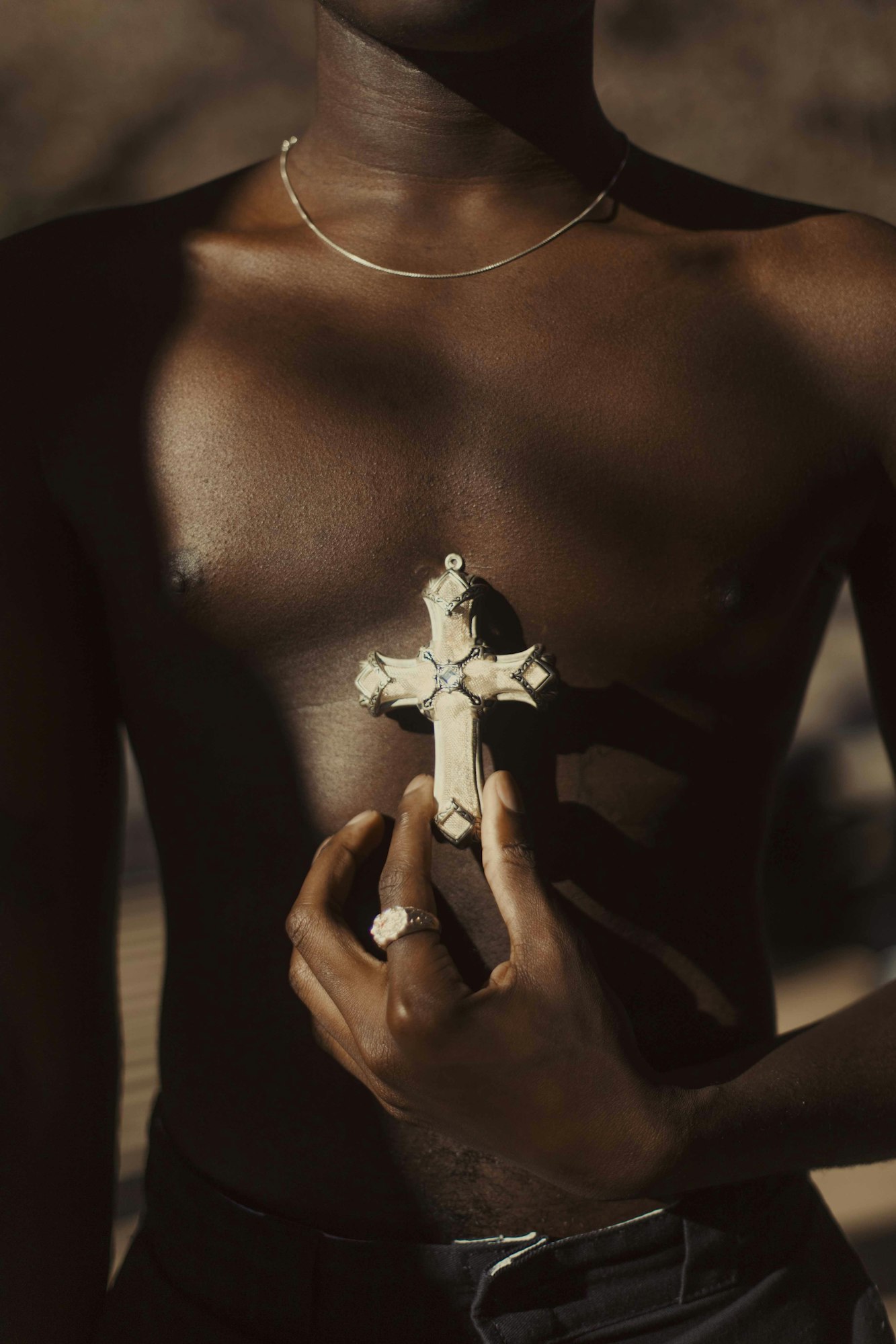 An upper body shot of a Black male holding a cross necklace, set against a blurred earth-tone background, captured with soft lighting.