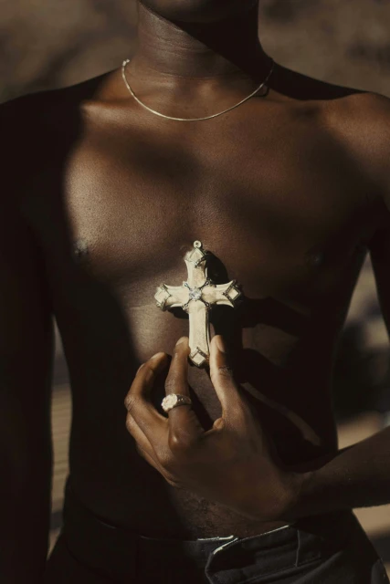 An upper body shot of a Black male holding a cross necklace, set against a blurred earth-tone background, captured with soft lighting.