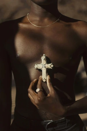 An upper body shot of a Black male holding a cross necklace, set against a blurred earth-tone background, captured with soft lighting.