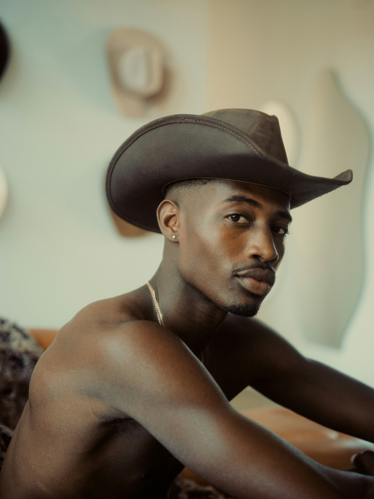 Portrait of a shirtless African-American man wearing a cowboy hat, looking at the camera, with cowboy hats on the background wall.