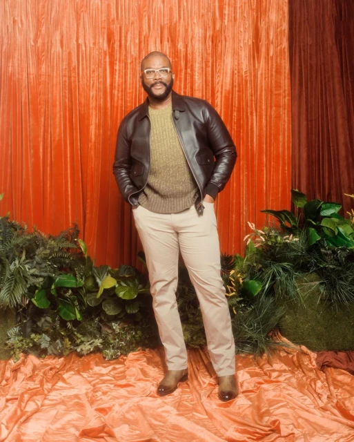 A full-length portrait of a Black man with glasses, in a brown jacket and tan pants, posed against an orange curtain and foliage backdrop.