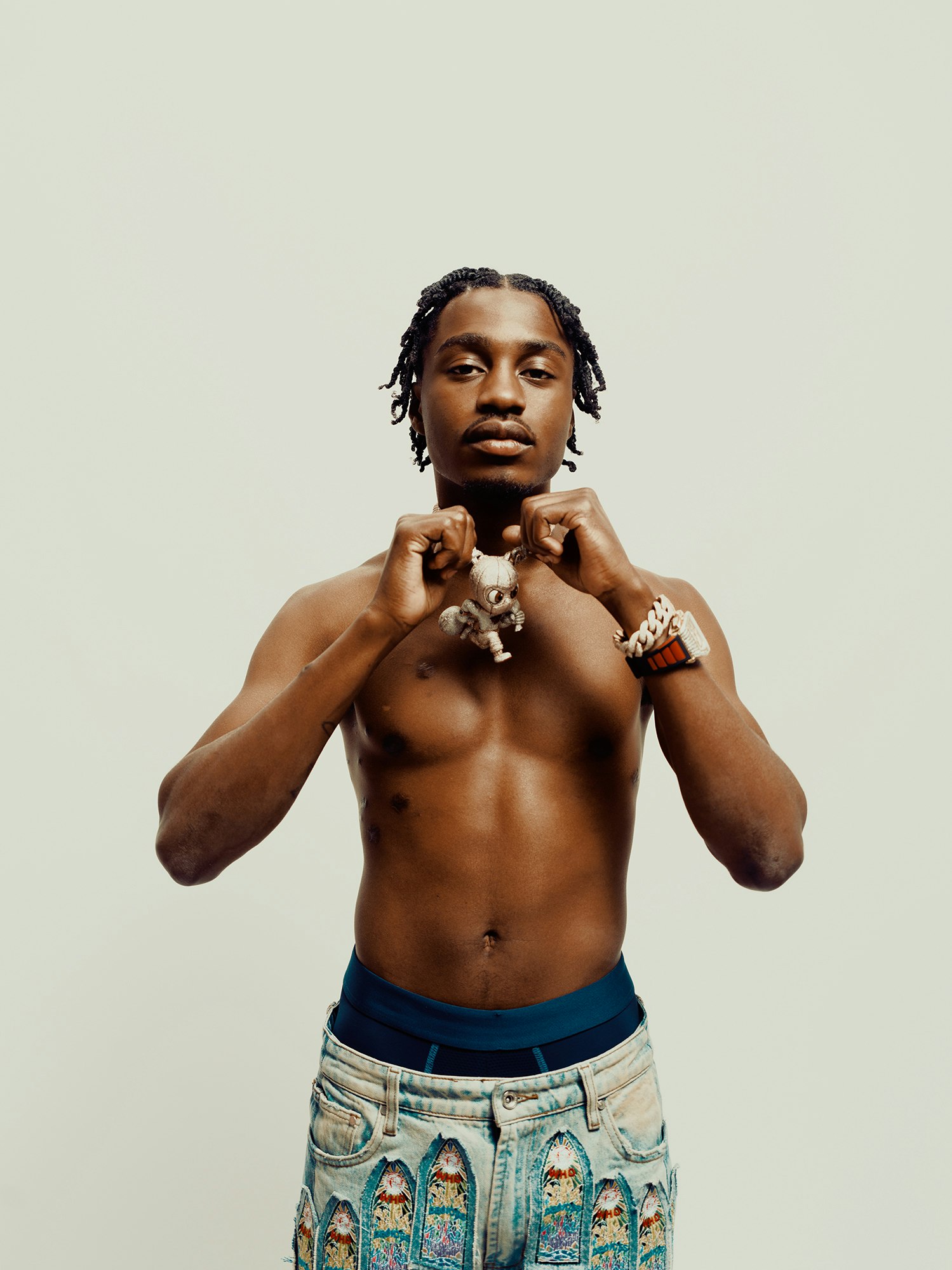 Portrait of shirtless adult male with braided hair, jewelry, and patterned jeans in a studio with a neutral background.