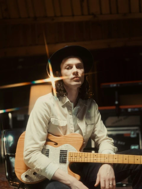 Portrait of a musician in a hat holding a light-colored electric guitar in an indoor setting with stage lighting effects.