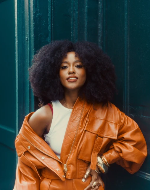 Portrait of an African-American woman with afro hairstyle wearing an orange leather jacket and matching pants posing against a green door.