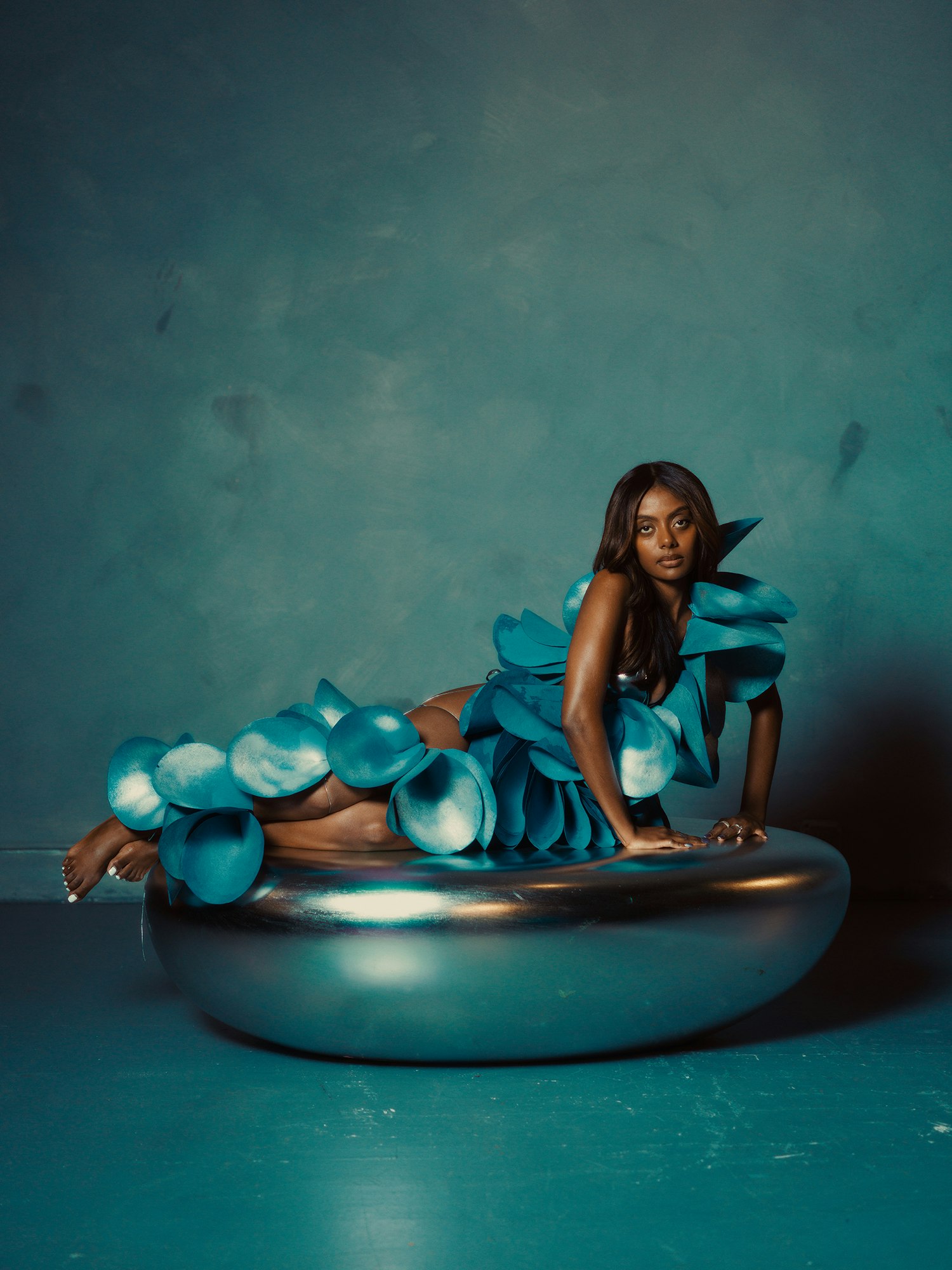 Adult female fashion model poses in an abstract blue floral dress on a silver bowl against a textured blue background in a studio.