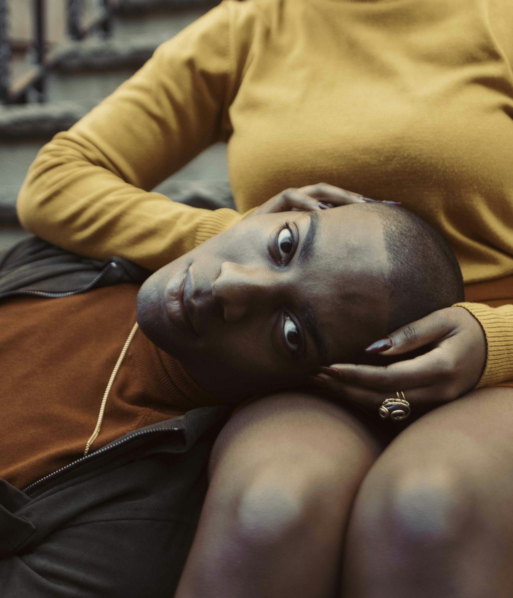 A close-up portrait of a young Black man resting his head in the lap of a woman wearing a mustard-yellow top. The shot is warm and intimate.
