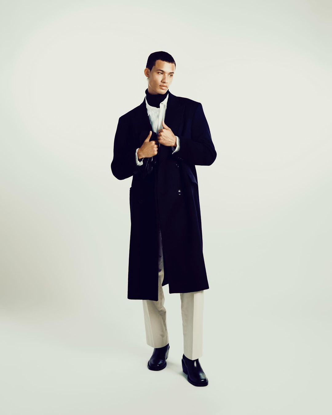 Model wearing a dark long coat over white shirt and light trousers against a neutral background
