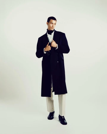 Model wearing a dark long coat over white shirt and light trousers against a neutral background