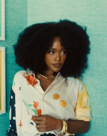 Woman with afro and patterned shirt poses in front of blue textured wall with framed artwork, showcasing fashion and portrait photography.