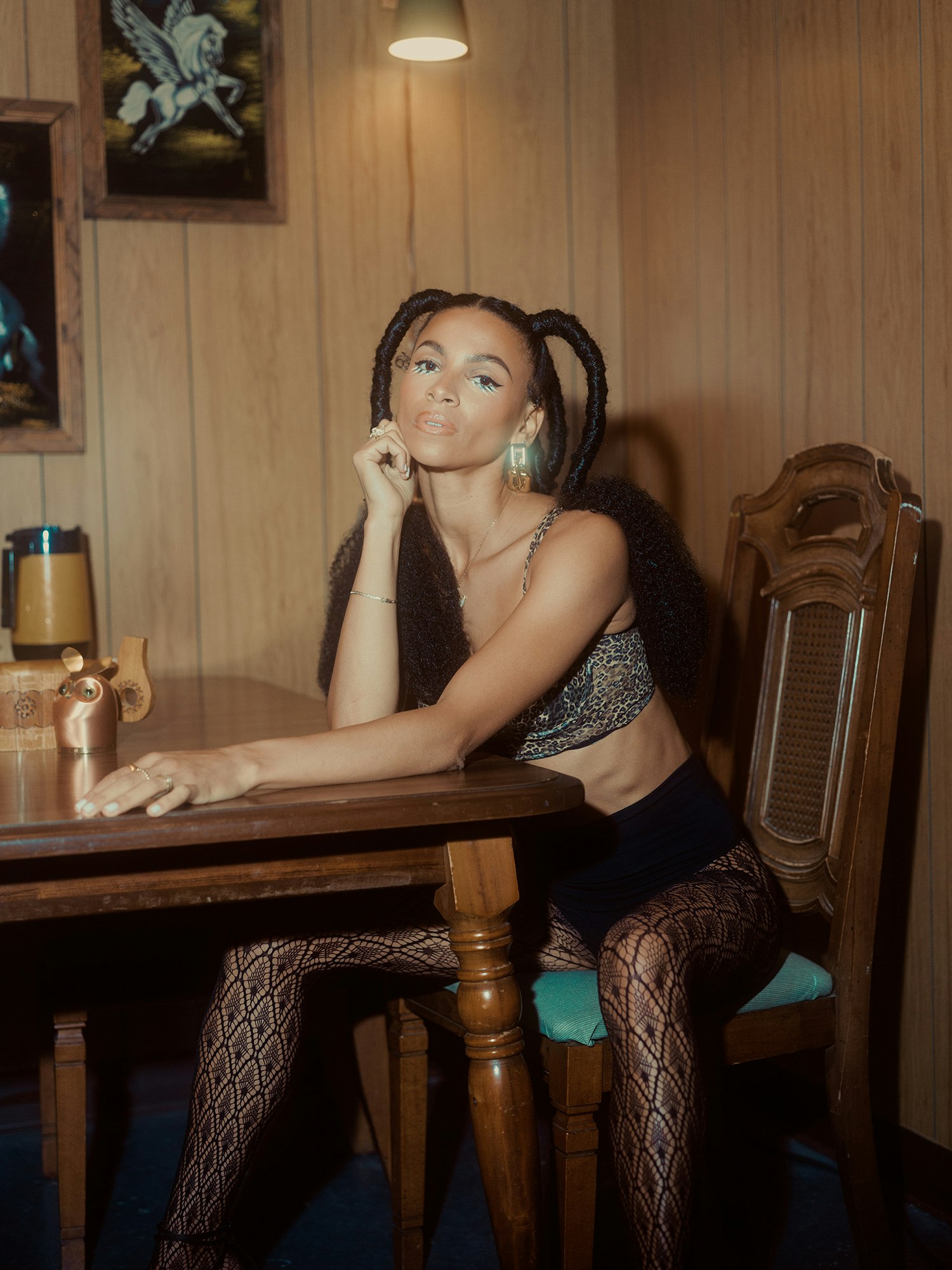 Portrait of a young woman with braided hair wearing a bra top and patterned tights, sitting at a wooden table in a wood-paneled room.