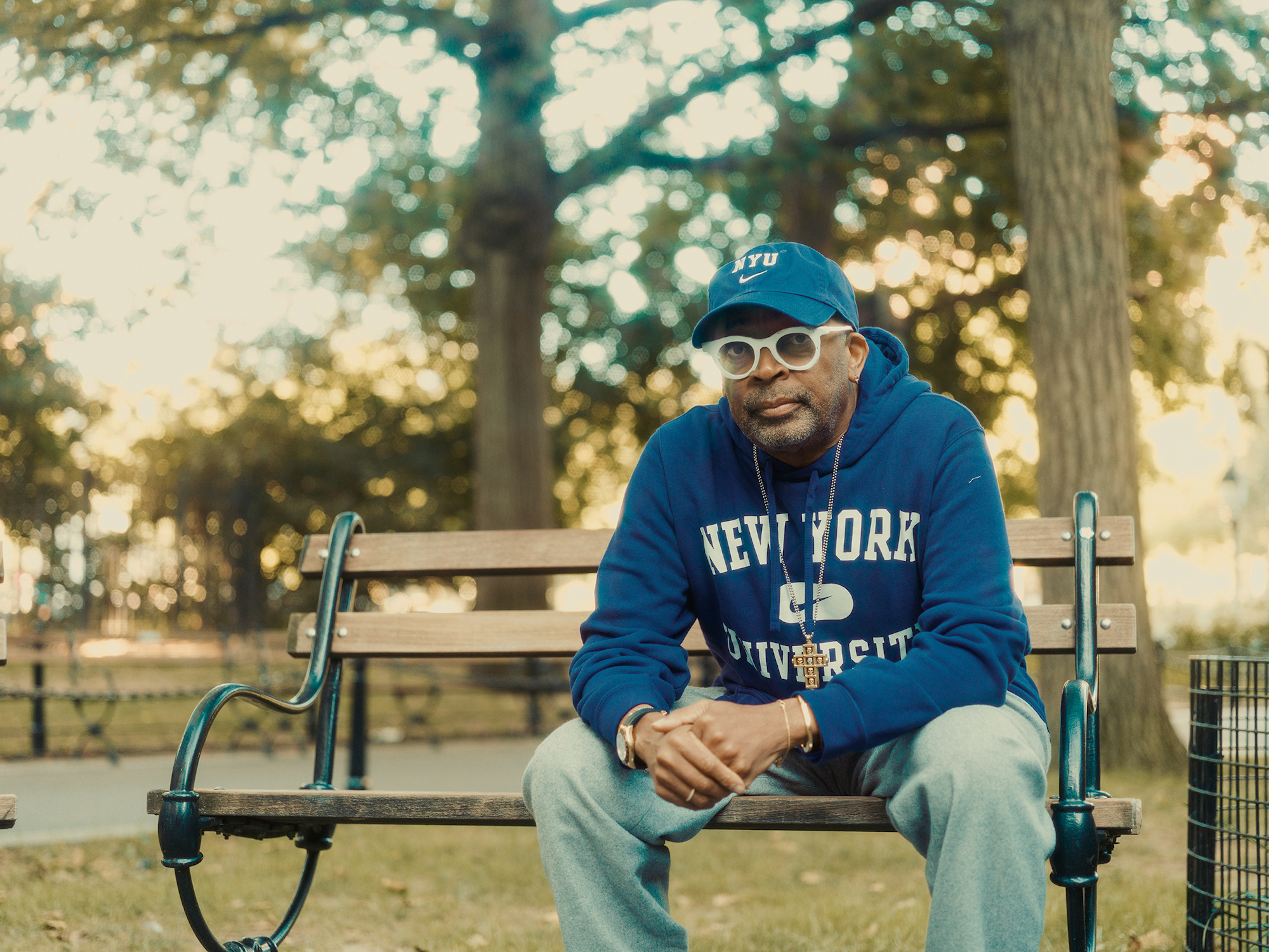 Adult male wearing a blue sweatshirt, glasses, and a baseball cap sits on a park bench surrounded by green trees and grass.