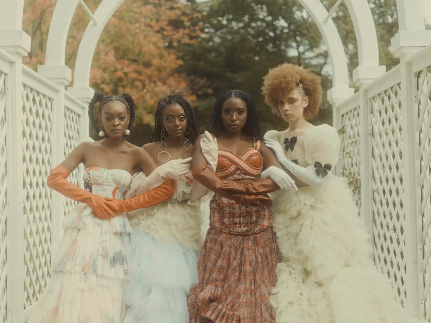 Four women in couture gowns pose under a white arbor with autumn foliage, creating a striking fashion editorial image.