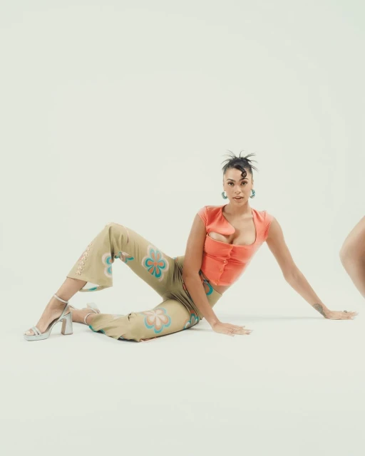 Adult woman in orange top and floral pants strikes a fashionable pose against a clean, white backdrop in a studio setting.
