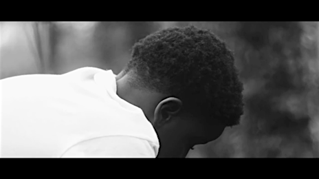 Monochrome close-up of a Black man with short hair, wearing a white shirt, head bowed in an outdoor environment with blurred background.