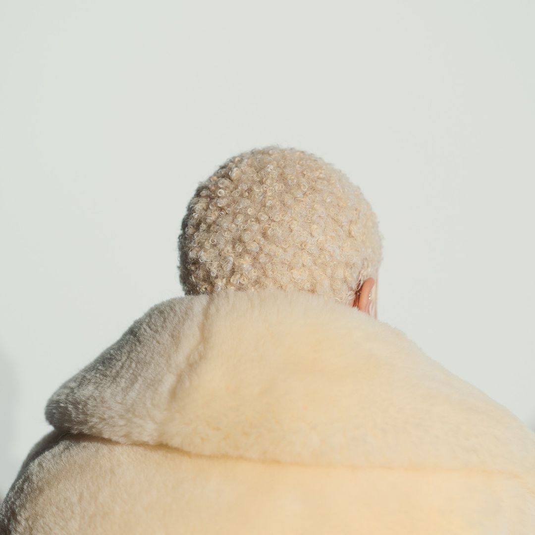 Back view of person with short, curly blonde hair wearing a light yellow, oversized fur coat against a stark white background.