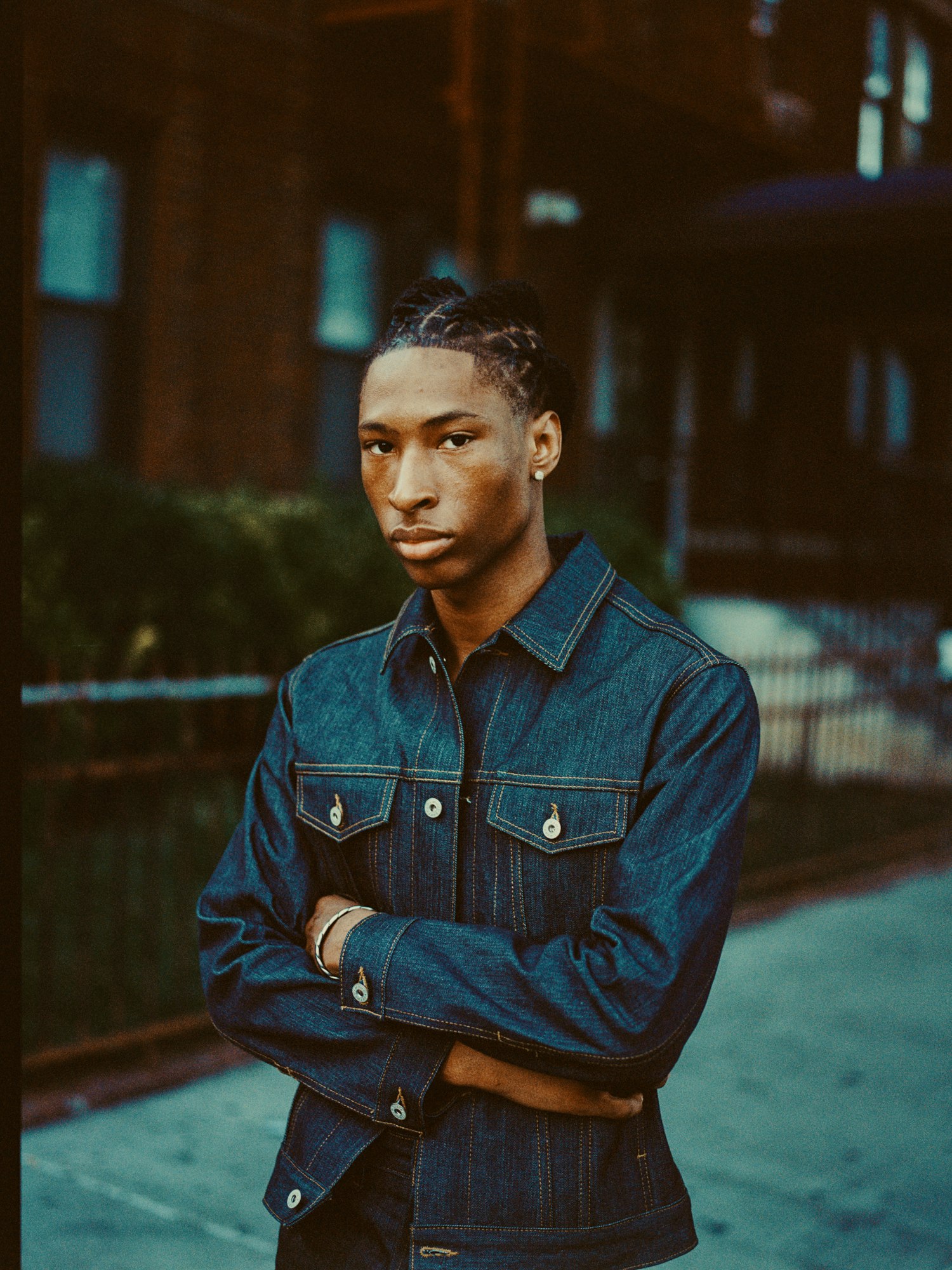 Portrait of a young adult Black man with braided hair, wearing a denim jacket with arms crossed, against an urban brick building backdrop.