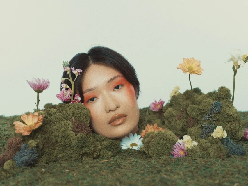 Eye-level medium shot of an Asian woman's head and shoulders surrounded by green moss and colorful flowers against a white background.