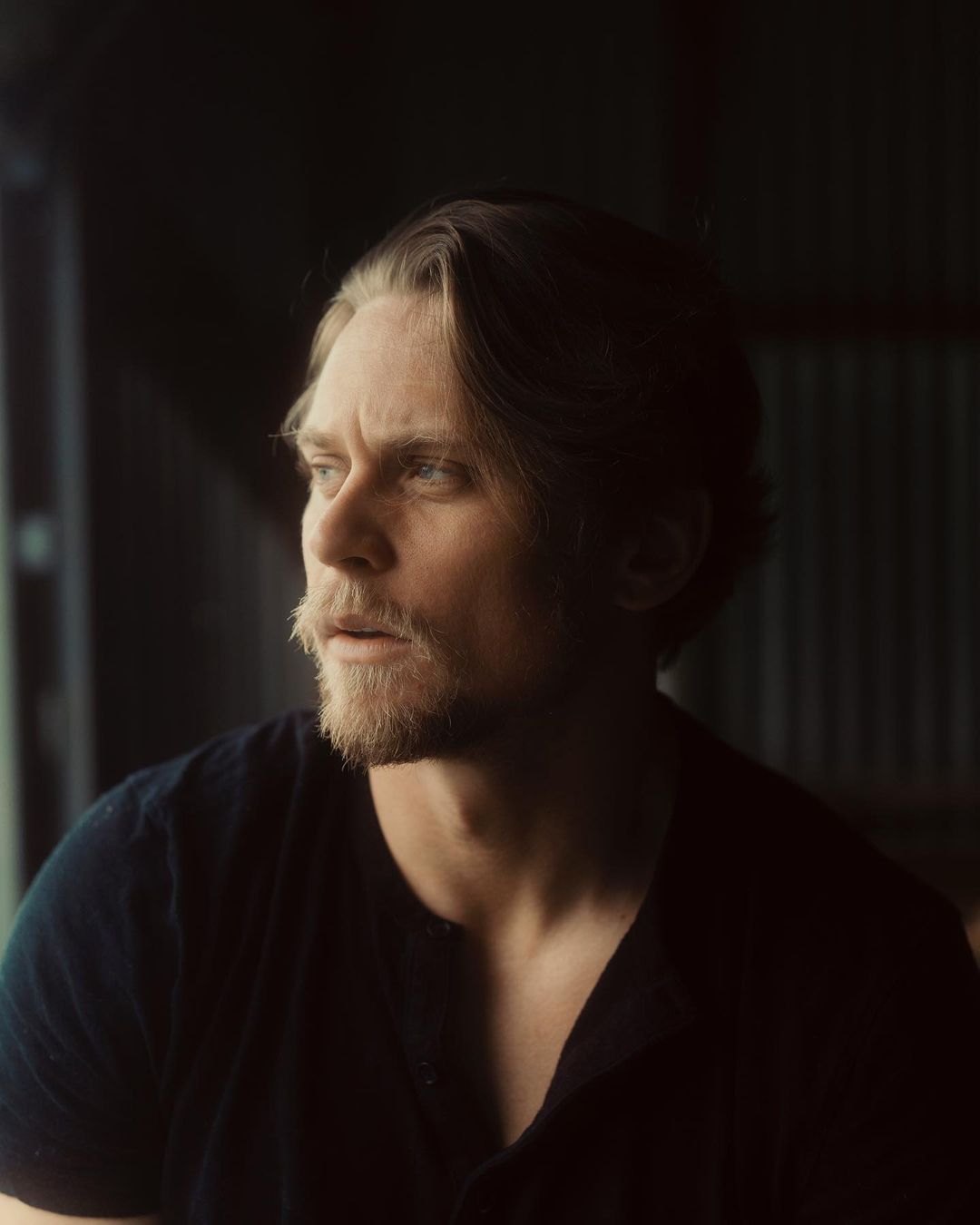 Portrait of an adult male with blond hair and a beard, wearing a dark shirt, looking out of frame with soft lighting in an indoor setting.