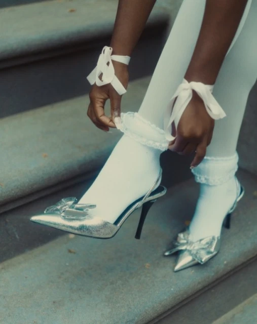 A person adjusts a white, frilled sock while wearing silver heels with bows, seated on grey stone steps with soft, diffused natural light.