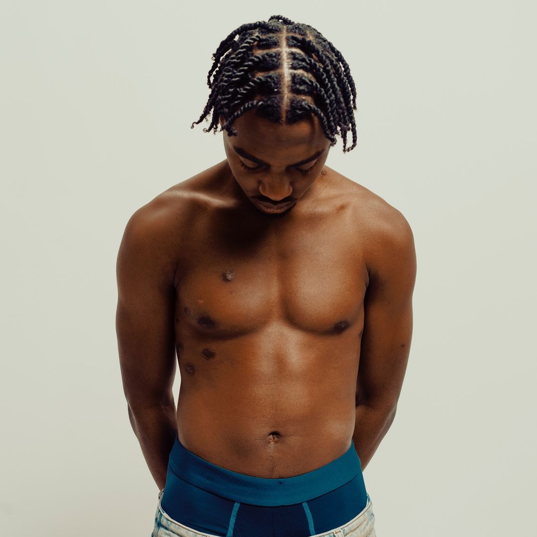Shirtless Black man with braided hair gazes down in a studio shot. The background is a soft off-white. He wears dark briefs.