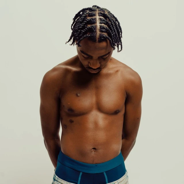 Shirtless Black man with braided hair gazes down in a studio shot. The background is a soft off-white. He wears dark briefs.