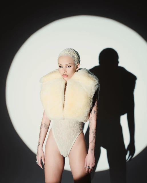 Woman with a shaved blonde haircut wears a textured bodysuit and a fuzzy cream-colored collar, posing in front of a spotlight.