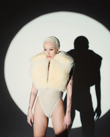 Woman with a shaved blonde haircut wears a textured bodysuit and a fuzzy cream-colored collar, posing in front of a spotlight.