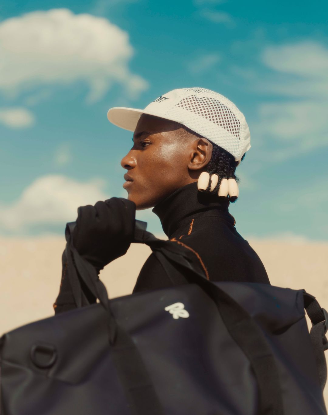 Profile portrait of a Black man wearing a white mesh cap and black duffel bag against a bright blue sky with clouds.