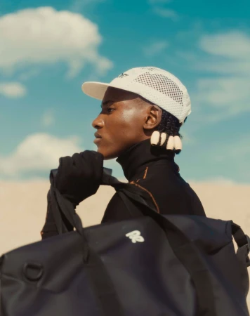 Profile portrait of a Black man wearing a white mesh cap and black duffel bag against a bright blue sky with clouds.