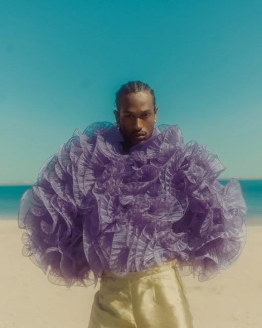 Man posing on beach wearing a purple ruffled top and gold pants. Clear turquoise sea and blue sky in the background.