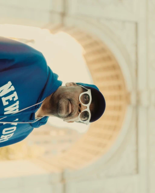 Adult male in blue hoodie and white sunglasses stands in front of an arch with golden hour lighting, creating a striking portrait.