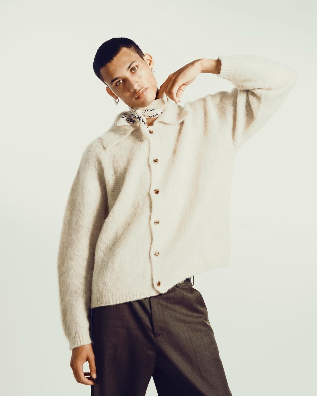 A man in a cream button-down cardigan and dark trousers posing against a plain background, photographed by Braylen Dion.