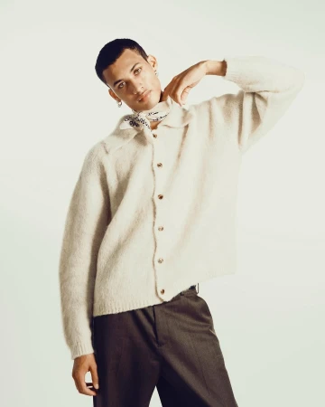 A man in a cream button-down cardigan and dark trousers posing against a plain background, photographed by Braylen Dion.