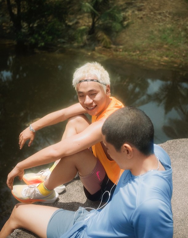 Two Asian men in athletic wear sit outdoors by a body of water, smiling in sunlight. One has blonde hair, and both wear running shoes.