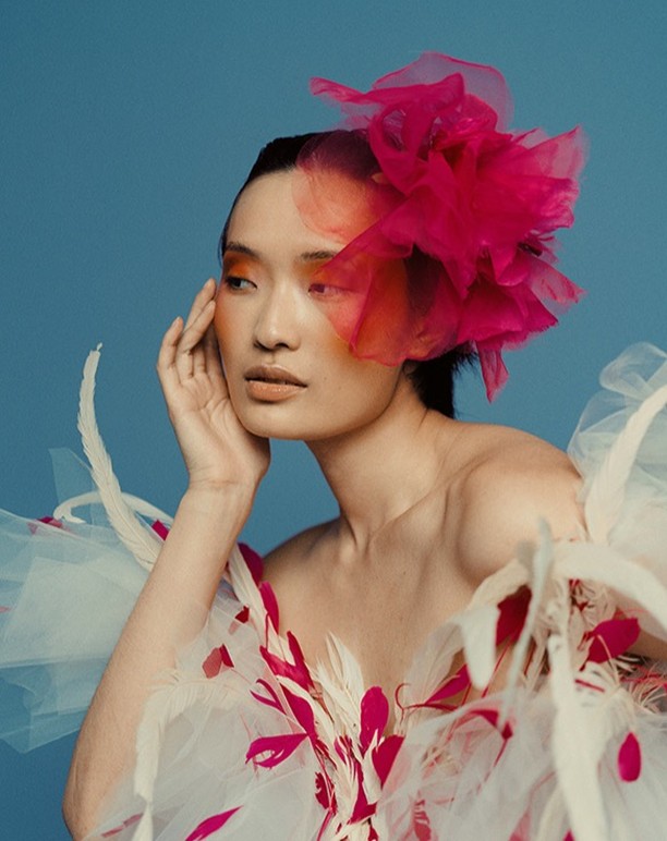 Portrait of an Asian woman with vibrant makeup and a large pink flower headpiece, posing in a white feathered dress against a blue backdrop.