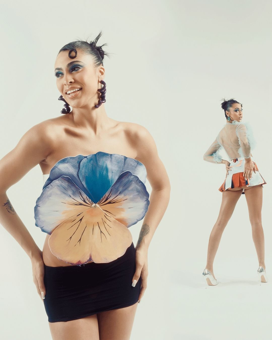 Two women strike poses in a white studio. One wears a flower top and black skirt, and the other wears a matching skirt set with heels.