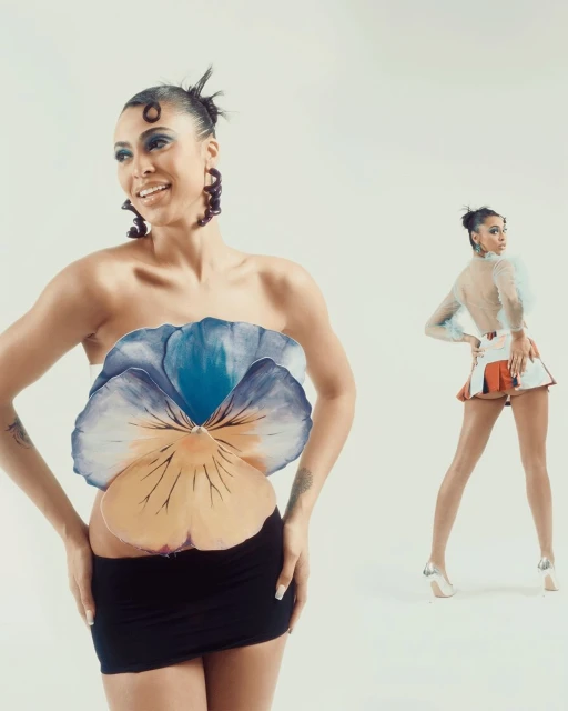 Two women strike poses in a white studio. One wears a flower top and black skirt, and the other wears a matching skirt set with heels.