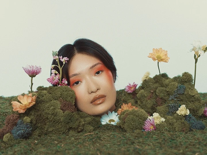 An Asian woman with short black hair and vibrant orange makeup surrounded by moss and flowers in a visually striking, surreal composition.