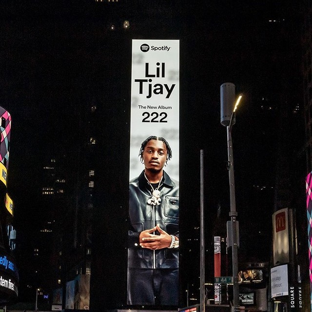 Billboard at night displays Lil Tjay album '222,' with the artist in a black jacket, against a backdrop of illuminated city buildings.