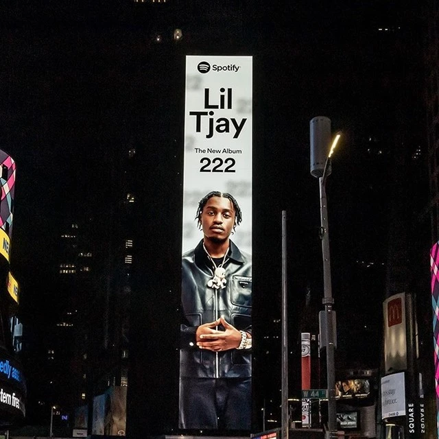 Billboard at night displays Lil Tjay album '222,' with the artist in a black jacket, against a backdrop of illuminated city buildings.