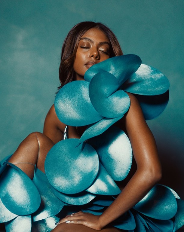 Elegant woman poses in an abstract, sculptural teal gown, eyes closed, against a mottled teal backdrop in a studio setting.