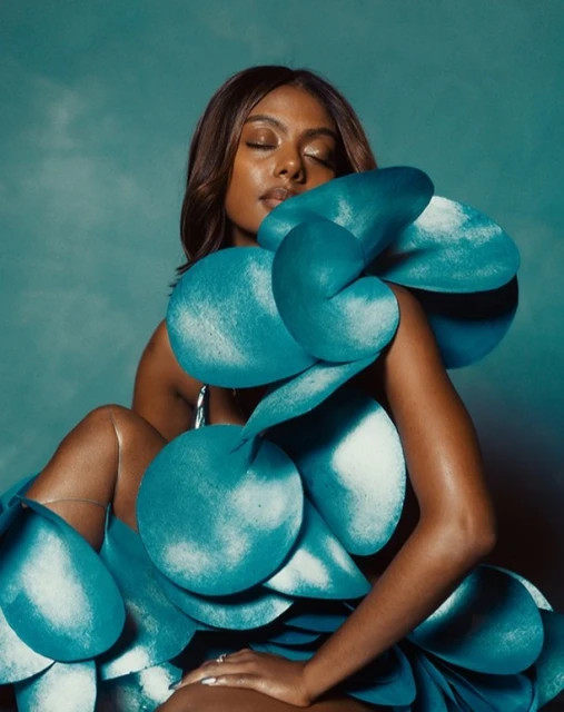 Elegant woman poses in an abstract, sculptural teal gown, eyes closed, against a mottled teal backdrop in a studio setting.