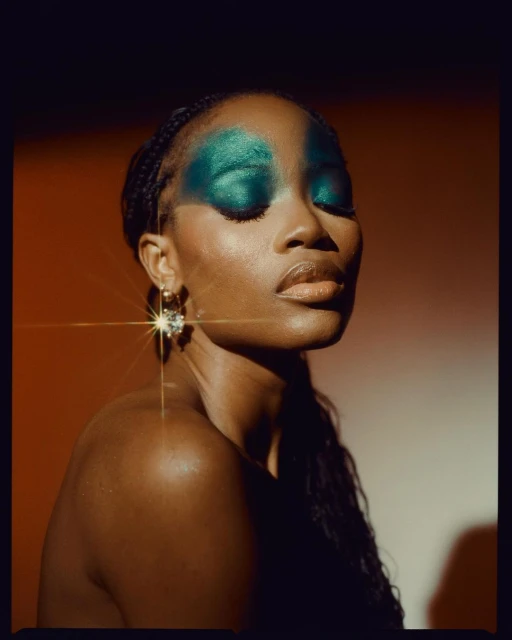 Portrait of a Black woman with braided hair, closed eyes, and vibrant teal eyeshadow, posing against a warm-toned background with a star filter.